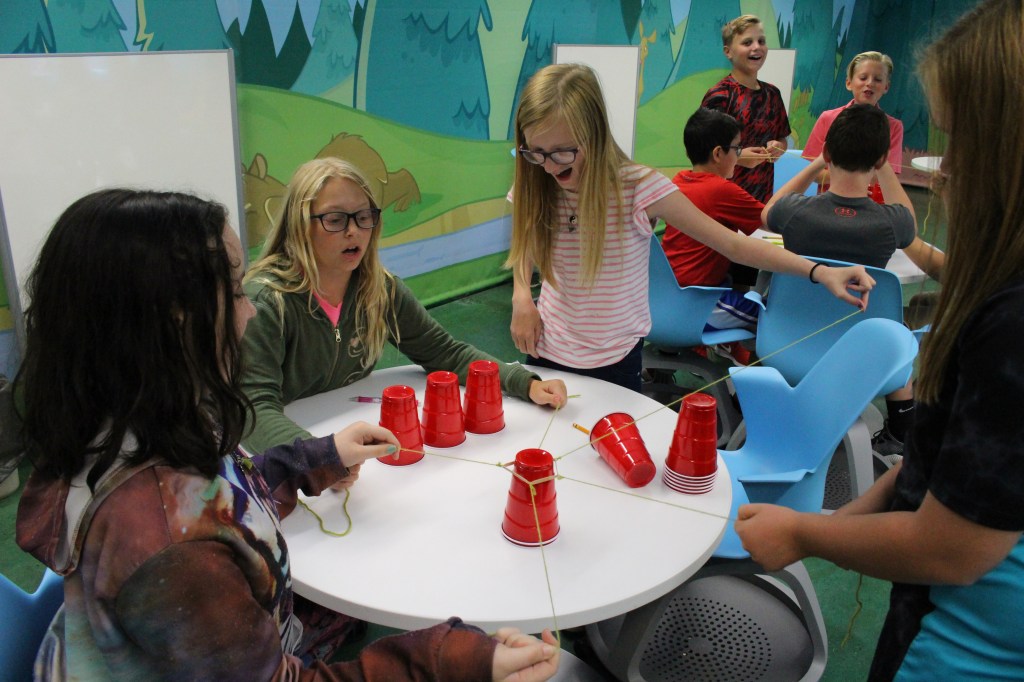 Students modeling the Four C's in a team-building activity by stacking cups using a "tool" made of a rubber band and yarn.